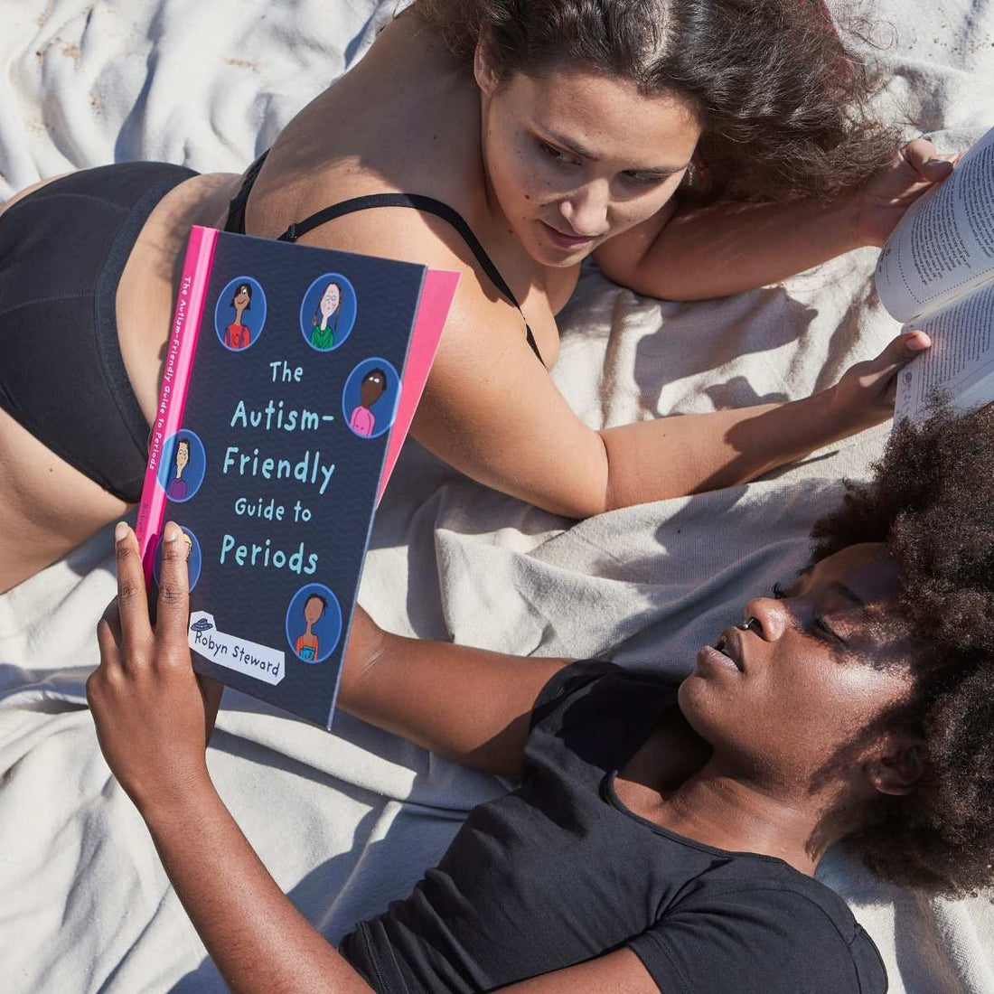 Two people relax on a blanket reading The Autism-Friendly Guide to Periods—a moment of connection, comfort, and inclusive period education under the sun.